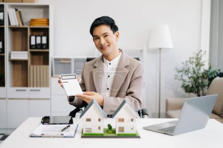 Photo for Young real estate agent worker working with laptop at table in modern office and small houses beside it - Royalty Free Image