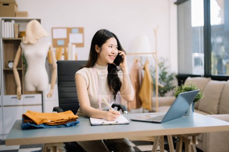 Téléchargez les photos : Femme tailleur asiatique travaillant sur des vêtements dans un atelier de couture. belle jeune créatrice de mode sourire après le succès dans le studio moderne - en image libre de droit
