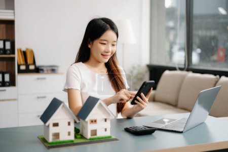 Photo for Young real estate agent worker working with laptop and smartphone at table in modern office and small houses model beside it - Royalty Free Image