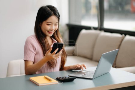 Téléchargez les photos : Asiatique fille bibliothèque assis au bureau en utilisant smartphone et ordinateur portable pour rechercher des informations en ligne. - en image libre de droit