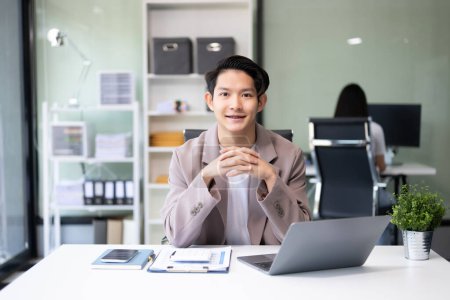 Foto de Joven atractivo asiático trabajador de oficina en trajes de negocios sonriendo a cámara en la oficina - Imagen libre de derechos