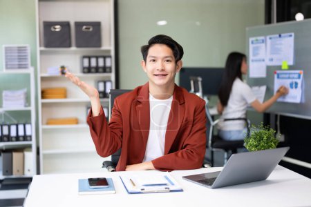 Foto de Joven atractivo asiático trabajador de oficina en trajes de negocios sonriendo a la cámara en la oficina y haciendo gestos de manos - Imagen libre de derechos