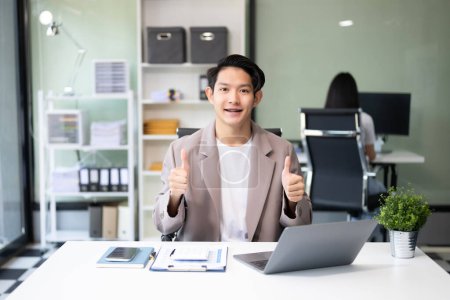 Foto de Joven atractivo asiático trabajador de oficina en trajes de negocios sonriendo a la cámara en la oficina y mostrando los pulgares hacia arriba - Imagen libre de derechos