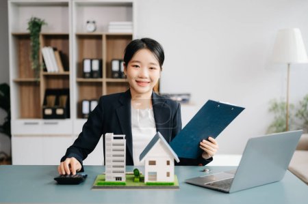 Foto de Trabajador joven agente de bienes raíces que trabaja con el ordenador portátil en la mesa en la oficina moderna y el modelo de casas pequeñas junto a ella - Imagen libre de derechos