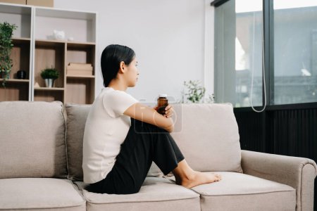 Téléchargez les photos : Asiatique femme sentiment dépression assis sur le canapé avec bouteille de pilules dans la maison - en image libre de droit