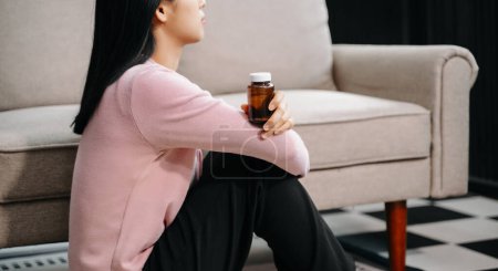 Téléchargez les photos : Femme asiatique se sentant dépression assis sur le sol au canapé et tenant bouteille de pilules dans la maison - en image libre de droit