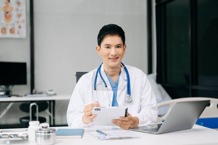 Téléchargez les photos : Médecin asiatique assis sur la table à l'hôpital de bureau. Masculin thérapeute utilisant tablette numérique et ordinateur portable pour le travail - en image libre de droit