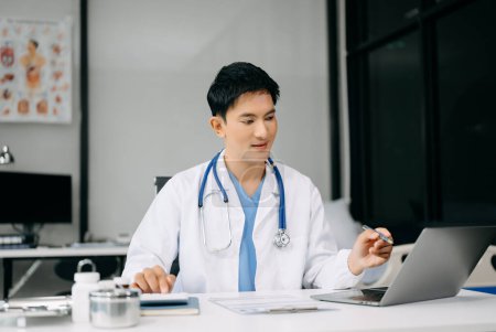 Foto de Médico asiático sentado en la mesa en el hospital de la oficina. Hombre terapeuta usando portátil para el trabajo - Imagen libre de derechos