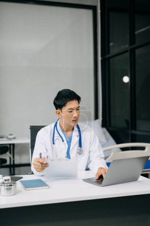 Foto de Médico asiático sentado en la mesa en el hospital de la oficina. Hombre terapeuta usando portátil para el trabajo - Imagen libre de derechos