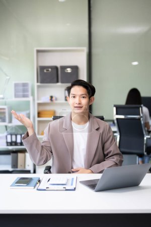 Foto de Joven atractivo asiático trabajador de oficina en trajes de negocios sonriendo a la cámara en la oficina y haciendo gestos de manos - Imagen libre de derechos