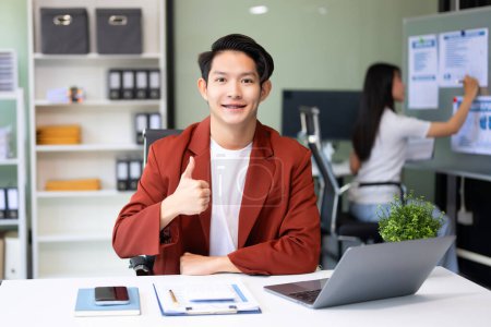 Foto de Joven atractivo asiático trabajador de oficina en trajes de negocios sonriendo a la cámara en la oficina y mostrando los pulgares hacia arriba - Imagen libre de derechos