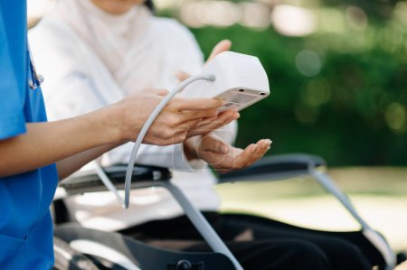 Anciano asiático senior mujer en silla de ruedas con asiático cuidadoso cuidador que mide la presión arterial en el jardín. 