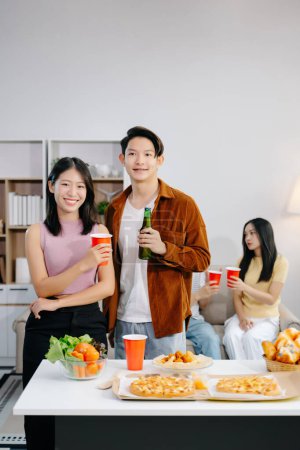 Téléchargez les photos : Groupe de jeunes amis asiatiques ayant, profiter de soirée ensemble à la maison. Jeunes hommes et femmes attrayants s'amusant, prenant des boissons, célébrant à la maison moderne - en image libre de droit