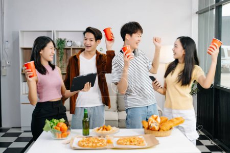 Foto de Grupo de jóvenes amigos asiáticos teniendo, disfrutar de la fiesta nocturna juntos en casa. Atractivos hombres y mujeres jóvenes que se divierten mientras usan teléfonos inteligentes y tabletas en el hogar moderno - Imagen libre de derechos