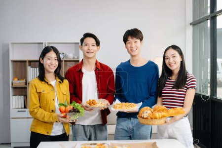 Téléchargez les photos : Groupe de jeunes amis asiatiques ayant, profiter de soirée ensemble à la maison. Jeunes hommes et femmes attirants tenant de la nourriture dans les assiettes et célébrant à la maison moderne - en image libre de droit
