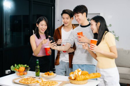 Photo for Group of Young Asian friends having, enjoy evening party together at home. Attractive young men and women having fun using smartphone and  celebrating at modern home - Royalty Free Image