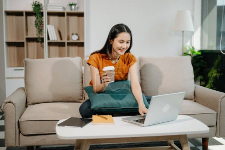Photo for Young asian woman relaxing on sofa at home and using laptop computer while drinking coffee - Royalty Free Image