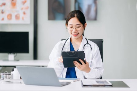 Une jeune femme asiatique confiante en uniforme médical blanc est assise au bureau et travaille sur ordinateur. Souriant utilisation ordinateur portable écrire dans un journal médical en clinique.
