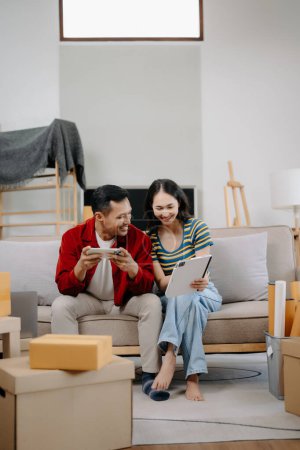 Foto de Asiático joven pareja atractiva hombre y mujer utilizan tableta y teléfono inteligente para muebles de compras en línea, para decorar la nueva casa. joven casado asiático mudanza en nuevo hogar - Imagen libre de derechos