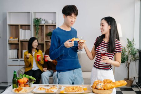 Foto de Grupo de jóvenes amigos asiáticos teniendo, disfrutar de la fiesta nocturna juntos en casa. Atractivos hombres y mujeres jóvenes divirtiéndose, comiendo y disfrutando de bebidas, celebrando en el hogar moderno - Imagen libre de derechos