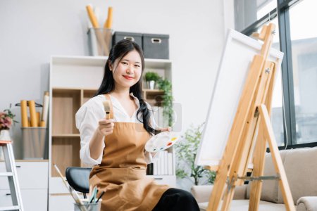 Photo for Asian Female painter doing artwork in art workshop, using painting supplies, oil pastels and posing at the camera creative space in art studio - Royalty Free Image