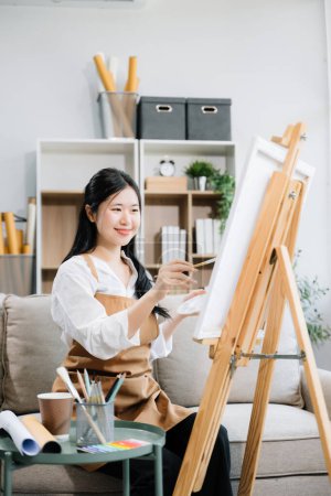 Photo for Asian Female painter doing artwork in art workshop, using painting supplies, oil pastels, creative space in art studio - Royalty Free Image