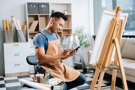 Photo for Asian male painter doing artwork in art workshop and using smartphone, painting supplies, oil pastels, canvas easel, creative space in art studio - Royalty Free Image