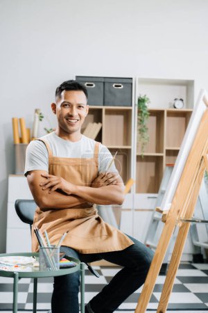 Photo for Asian male painter doing artwork in art workshop and posing at the camera, painting supplies, oil pastels, canvas easel, creative space in art studio - Royalty Free Image