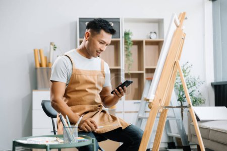Téléchargez les photos : Peintre asiatique homme faisant des œuvres d'art dans l'atelier d'art et en utilisant smartphone, fournitures de peinture, pastels à l'huile, chevalet en toile, espace créatif dans l'atelier d'art - en image libre de droit