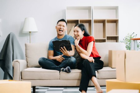 Happy asian young attractive couple man and woman with big boxes moving into a new house and using digital tablet 