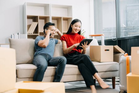 Happy asian young attractive couple man and woman with big boxes moving into a new house using digital tablet and smartphone 