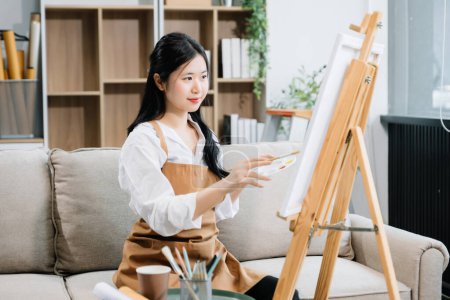 Photo for Asian Female painter doing artwork in art workshop, using painting supplies, oil pastels, creative space in art studio - Royalty Free Image