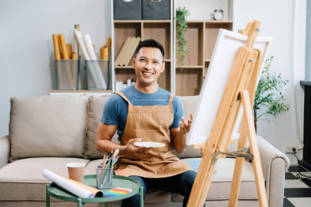 Photo for Asian male painter doing artwork in art workshop and posing at the camera, painting supplies, oil pastels, canvas easel, creative space in art studio - Royalty Free Image