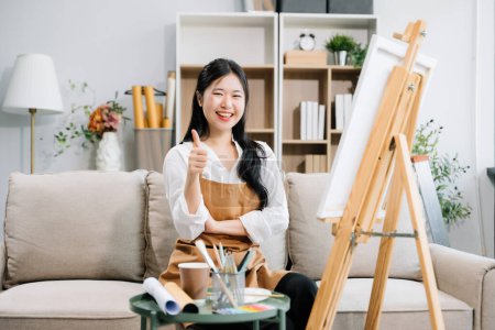 Photo for Asian Female painter doing artwork in art workshop, using painting supplies, oil pastels and showing thumbs up sign, creative space in art studio - Royalty Free Image