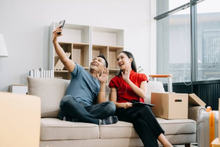 Happy asian young attractive couple man and woman with big boxes moving into a new house and taking selfie on smartphone 