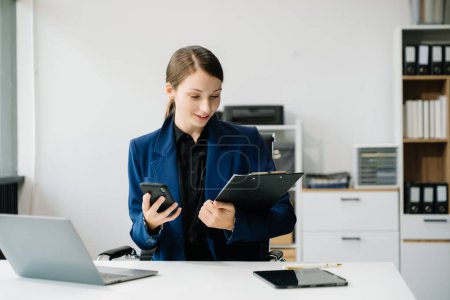 Téléchargez les photos : Femme d'affaires assise à un bureau en utilisant un smartphone et un ordinateur portable dans le bureau - en image libre de droit