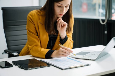 Photo for Businesswoman using laptop and writing in documents at desk in office - Royalty Free Image