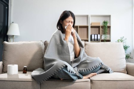 Junge Asiatin mit Grippesymptomen, mit einer Decke auf dem Sofa bedeckt und in Gewebe niesen. Konzept von Krankheit, Gesundheit und Genesung