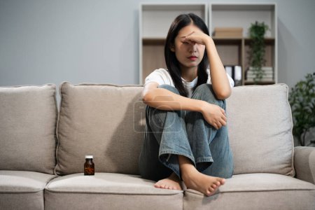 Téléchargez les photos : Asiatique femme dans la dépression assis sur canapé avec bouteille de pilules - en image libre de droit