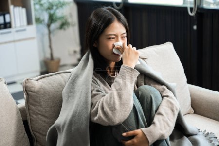 Young Asian woman suffering from flu symptoms, covered with a blanket on a sofa and sneezing in tissue. Concept of illness, healthcare and recovery