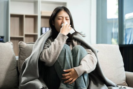 Foto de Mujer asiática joven que sufre de síntomas de gripe, cubierta con una manta en un sofá y estornudos en el tejido. Concepto de enfermedad, salud y recuperación - Imagen libre de derechos
