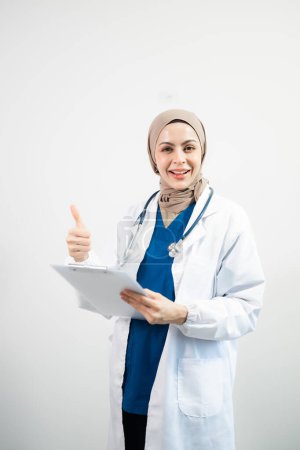 Muslimische arabische Ärztin im Hijab mit erhobenem Daumen auf isoliertem weißem Hintergrund im Studio