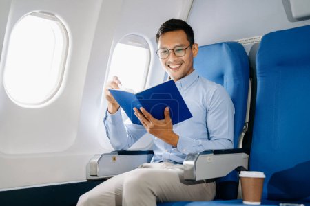 smiling Asian man sitting in a seat in airplane and reading book, going on a trip vacation. 