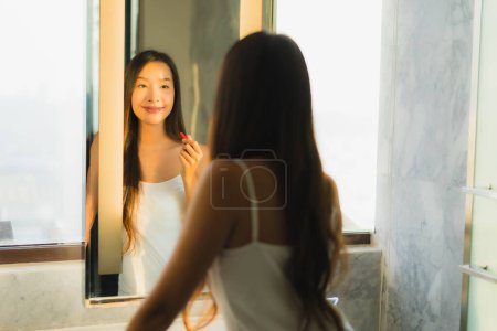 Téléchargez les photos : Portrait belle jeune femme asiatique maquillage avec de la poudre cosmétique et rouge à lèvres sur son visage facial dans la salle de bain intérieure - en image libre de droit