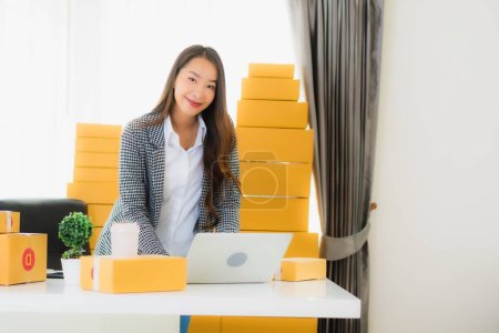 Foto de Retrato hermosa joven mujer de negocios asiática trabajo desde casa con teléfono móvil portátil con caja de cartón listo para el envío de compras en línea - Imagen libre de derechos