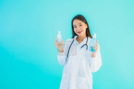 Téléchargez les photos : Portrait belle jeune femme asiatique médecin avec de l'alcool gel pour la main propre et protéger de covid19 ou coronavirus sur fond bleu isolé - en image libre de droit