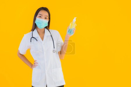 Téléchargez les photos : Portrait belle jeune asiatique médecin femme porter un masque avec gel d'alcool pour protéger de covid19 ou coronavirus sur fond isolé jaune - en image libre de droit