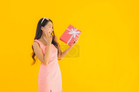 Téléchargez les photos : Portrait belle jeune femme asiatique avec boîte cadeau rouge sur fond jaune isolé - en image libre de droit