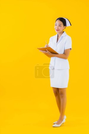 Téléchargez les photos : Portrait belle jeune femme asiatique thai infirmière montrer tableau blanc vide et travailler à la clinique ou à l'hôpital sur fond jaune isolé - en image libre de droit