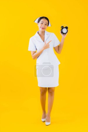 Photo for Portrait beautiful young asian woman thai nurse show clock or alarm on yellow isolated background - Royalty Free Image